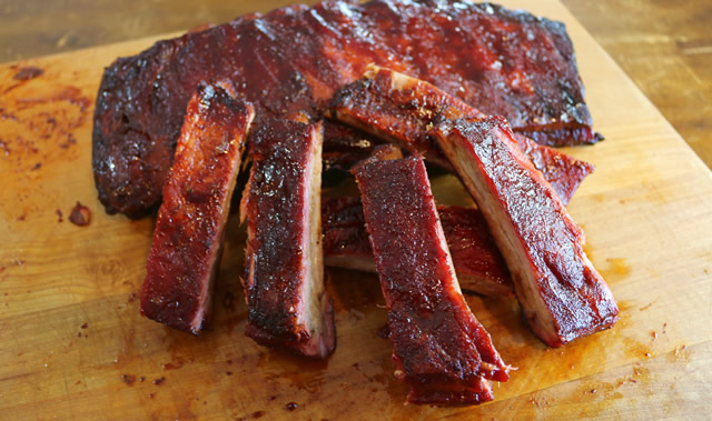 A rack of barbecued ribs cut into individual pieces on a wooden cutting board, deep red and brown charred exterior, rustic presentation.