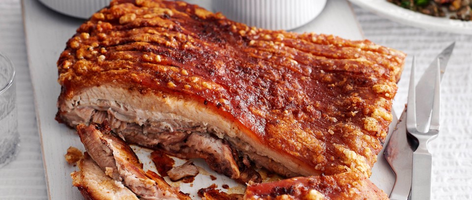 A sliced roast pork belly with crispy crackling and tender meat, served on a cutting board with a carving fork and knife.